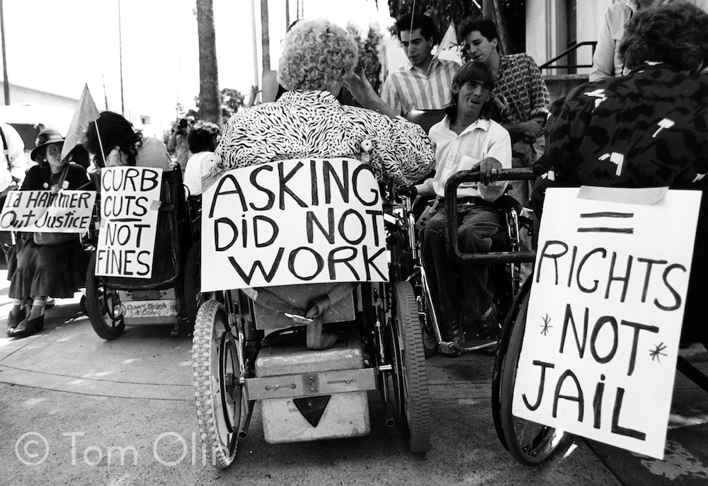 Black and white photo of the back of a person in a power wheelchair with a sign tacked on that reads "asking did not work," another person in a wheelchair beside has a sign that reads "equal rights not jail"