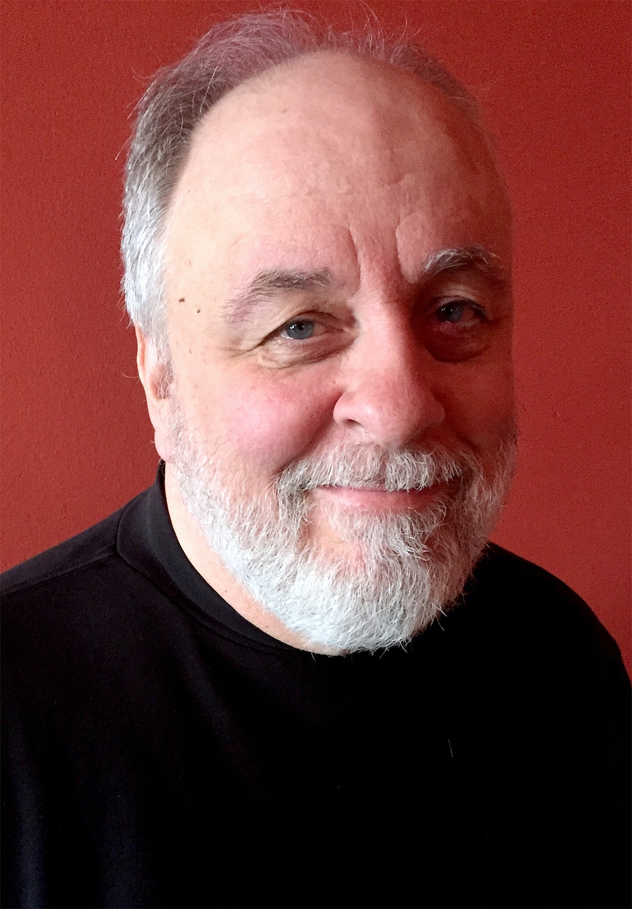 Portrait of Dan, an older white man with a beard. He's wearing a black turtle neck and a red background