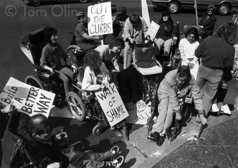 Black and white photo of mostly people in wheelchairs crowded around a street corner. One person in breaking the curb with cement dust around the object. People hold signs that read "cut the curbs," "walk of shame," and "build a better way"