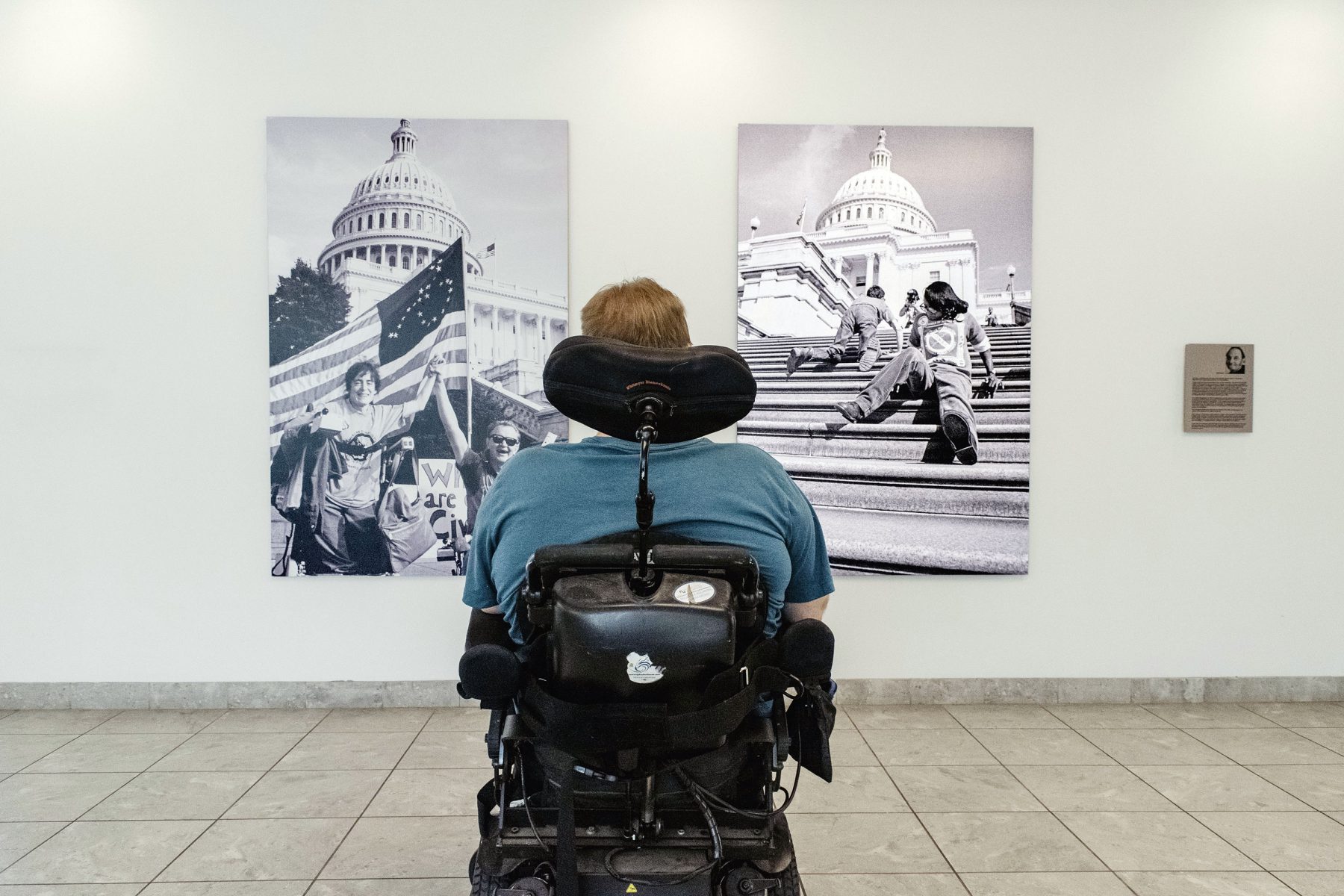 A person using a power chair faces away from the camera towards two large format Tom Olin Collection photos mounted on a wall