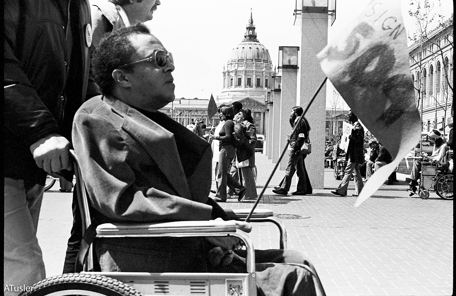 Black and white photo of profile view of a black man in sunglasses using manual wheelchair. A rotunda of a building is in the background.