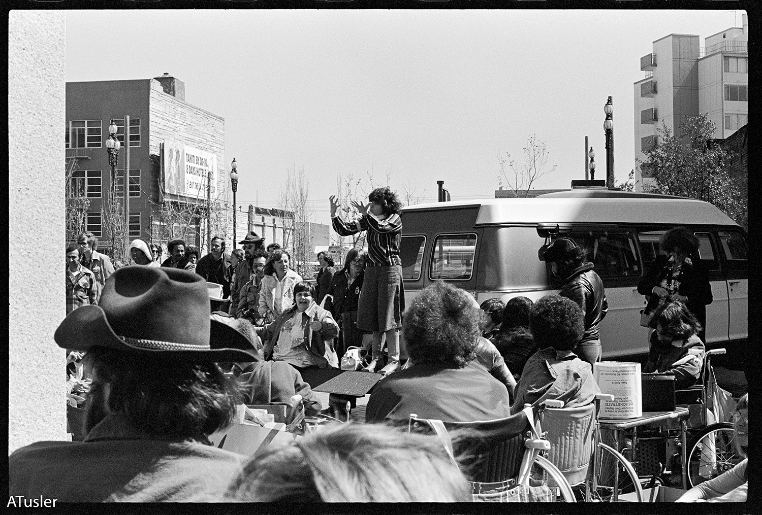 Black and white photo of crowd looking on, a person using ASL is seen at the back of a van's wheelchair lift