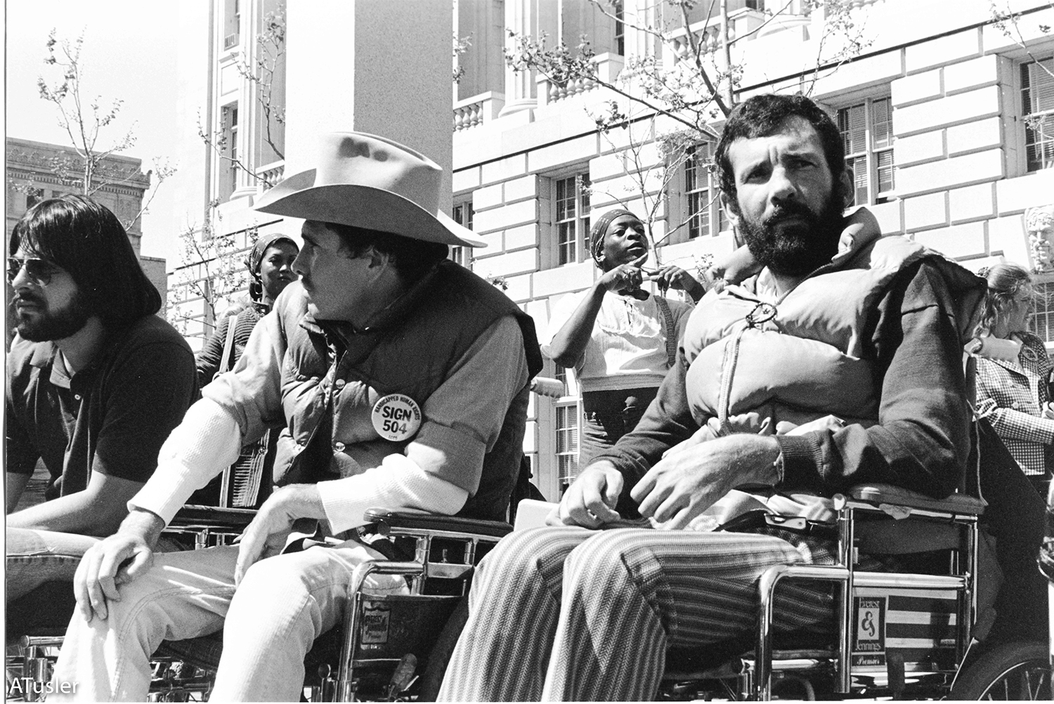Black and white photo of two men in manual wheelchairs, the exterior of the San Francisco federal building in the background