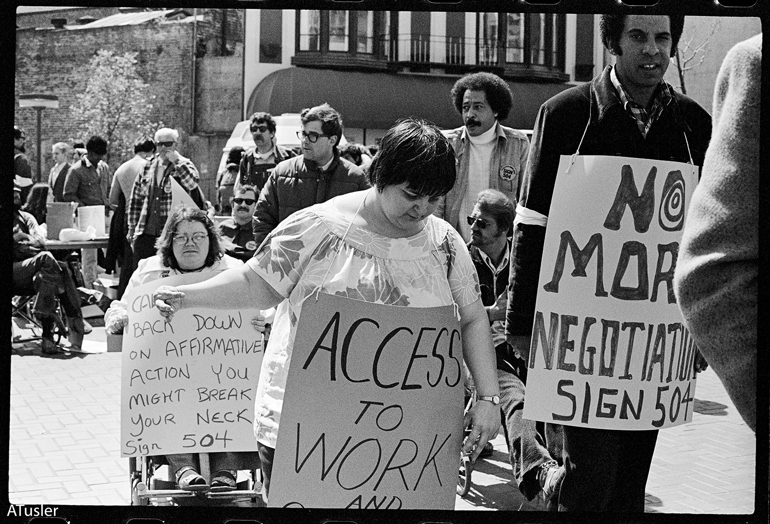 Black and white photo of people demonstrating with “No More Negotiations, Sign 504” and “Access to Work” hand-lettered signs.