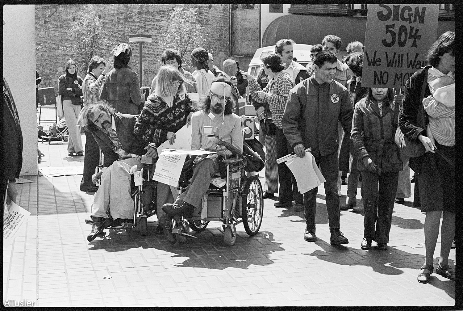 Black and white photo with two men in manual wheelhairs holding signs, other picketers off to the side