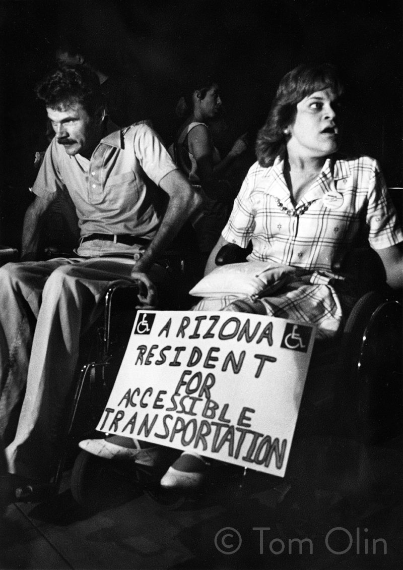Black and white photo of two people in wheelchairs. One is holding a sign that reads "Arizona Resident for Accessible Transportation"