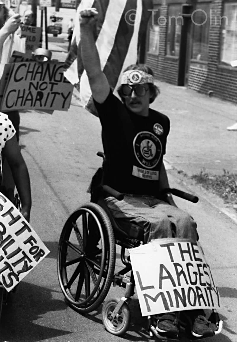 Black and white photo of person in a manual wheelchair holding up their fist. A sign resting on their legs reads "The Largest Minority," a posting in the back reads "Change not Charity"