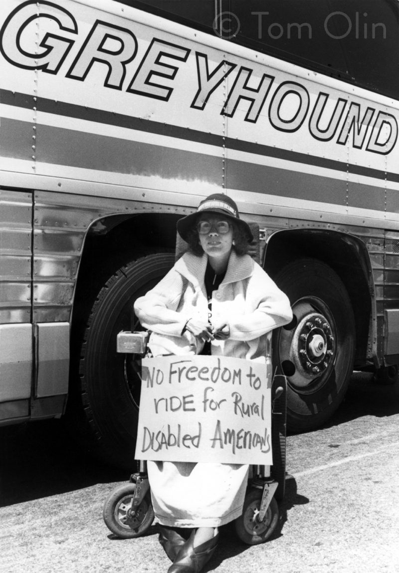 Black and white photo of person wearing a sun hat and wearing a long coat sits in a power wheelchair with a sign that reads "No freedom to ride for rural disabled americans."