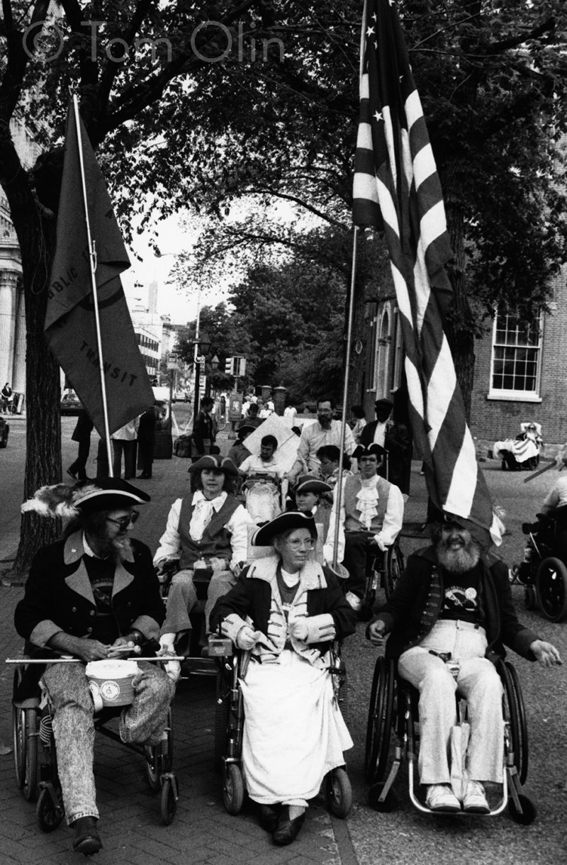 Black and white photo of three people in wheelchairs wearing period clothing and holding a U.S. flag, one with stars in the shape of the access symbol.