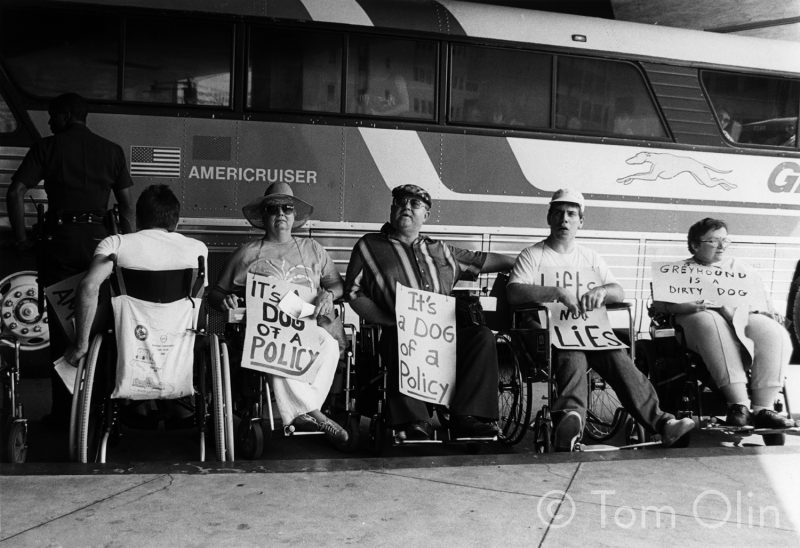 Black and white photo of people in front of a Greyhound bus, the people are in manual wheelchairs, holding signs that read "it's a dog of a policy," "lifts not lies," and "Greyhound is a dirty dog"