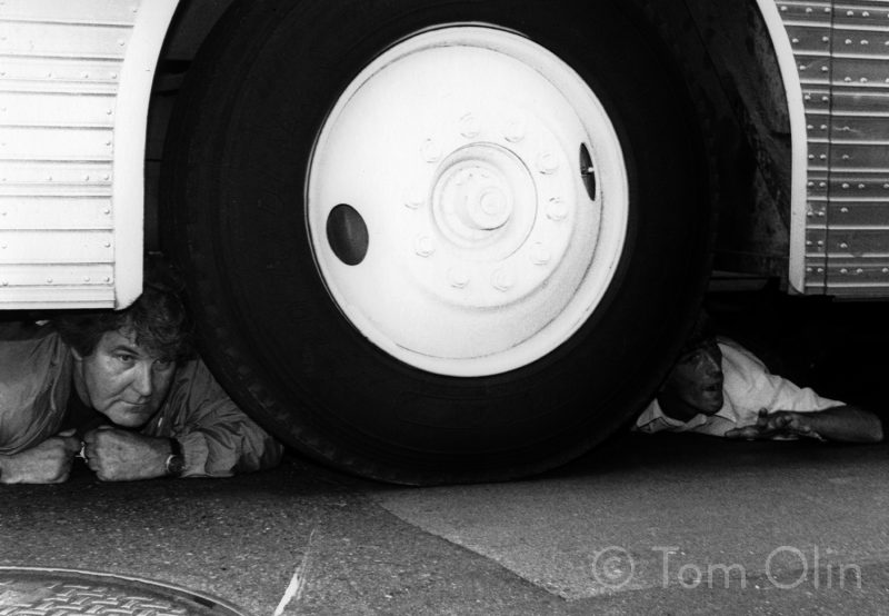 Black and white photo of a close up of a wheel on a but, there are two people underneath the bus, perched on each side of the large tire.