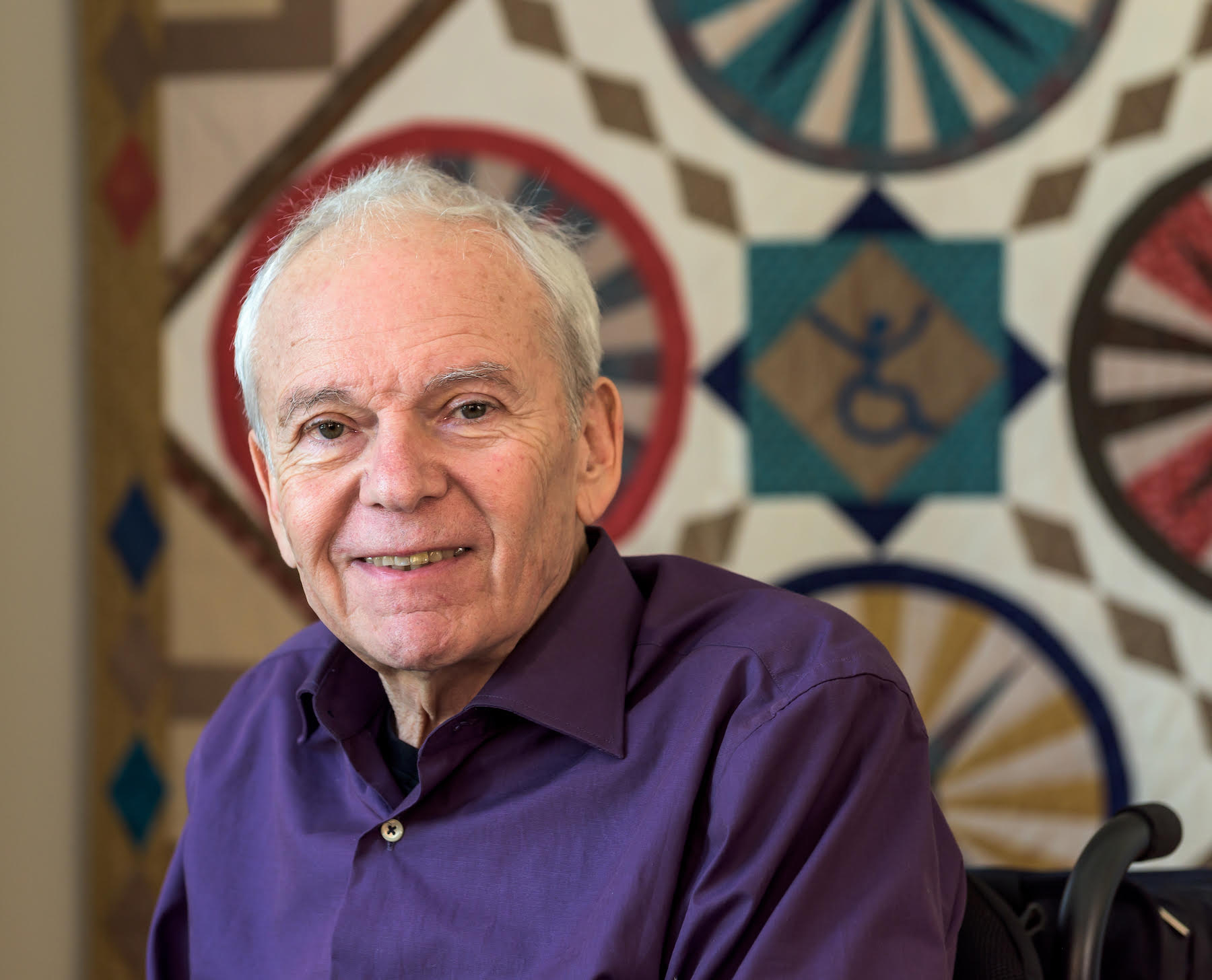 Portrait of Anthony, a white male in a purple collared shirt, in front of a quilt that has a the ADAPT logo on it.