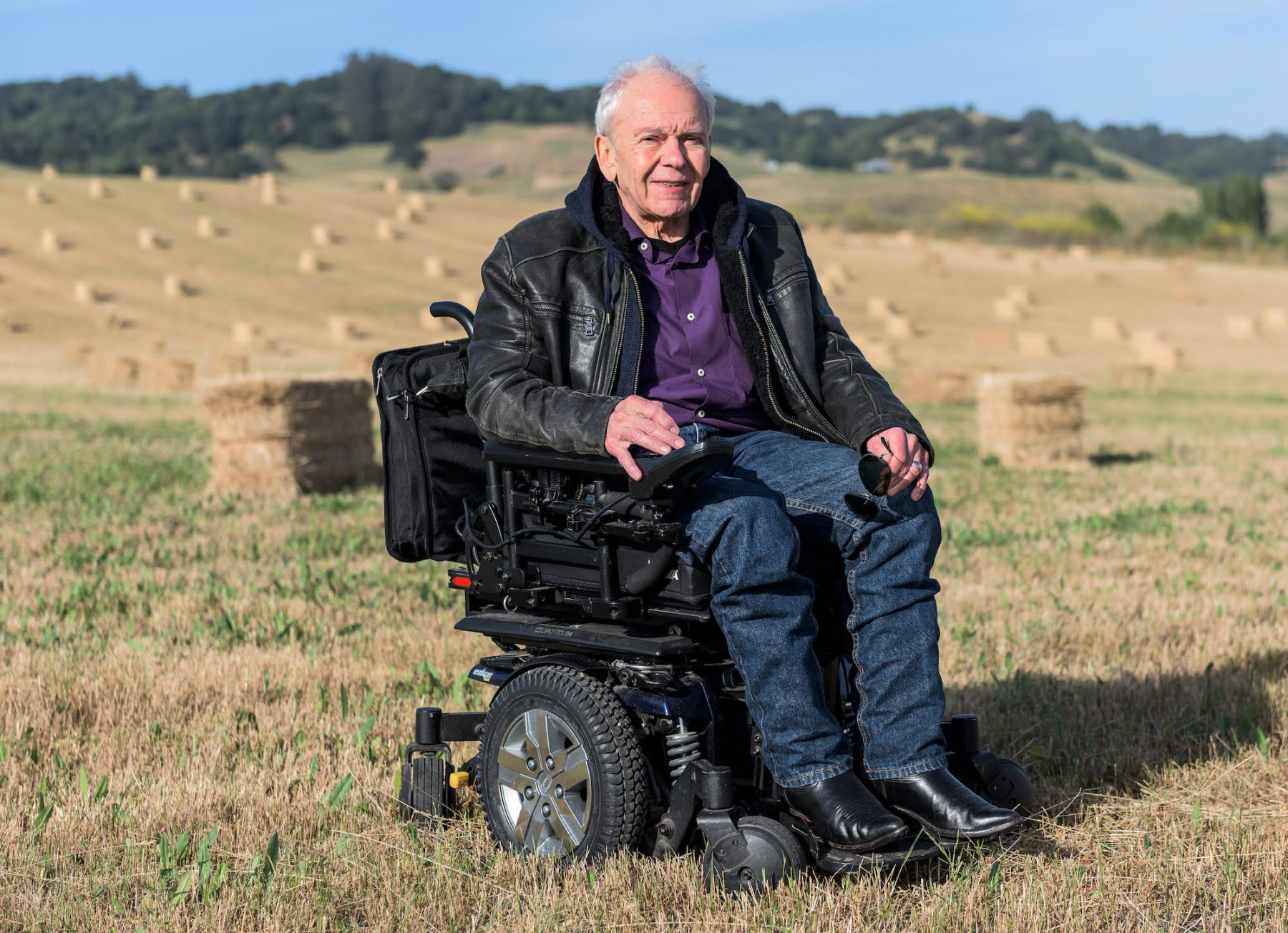 Anthony, a white male in a purple collared shirt, black leather jacket and jeans, sits in a power chair in a middle of a hay field.